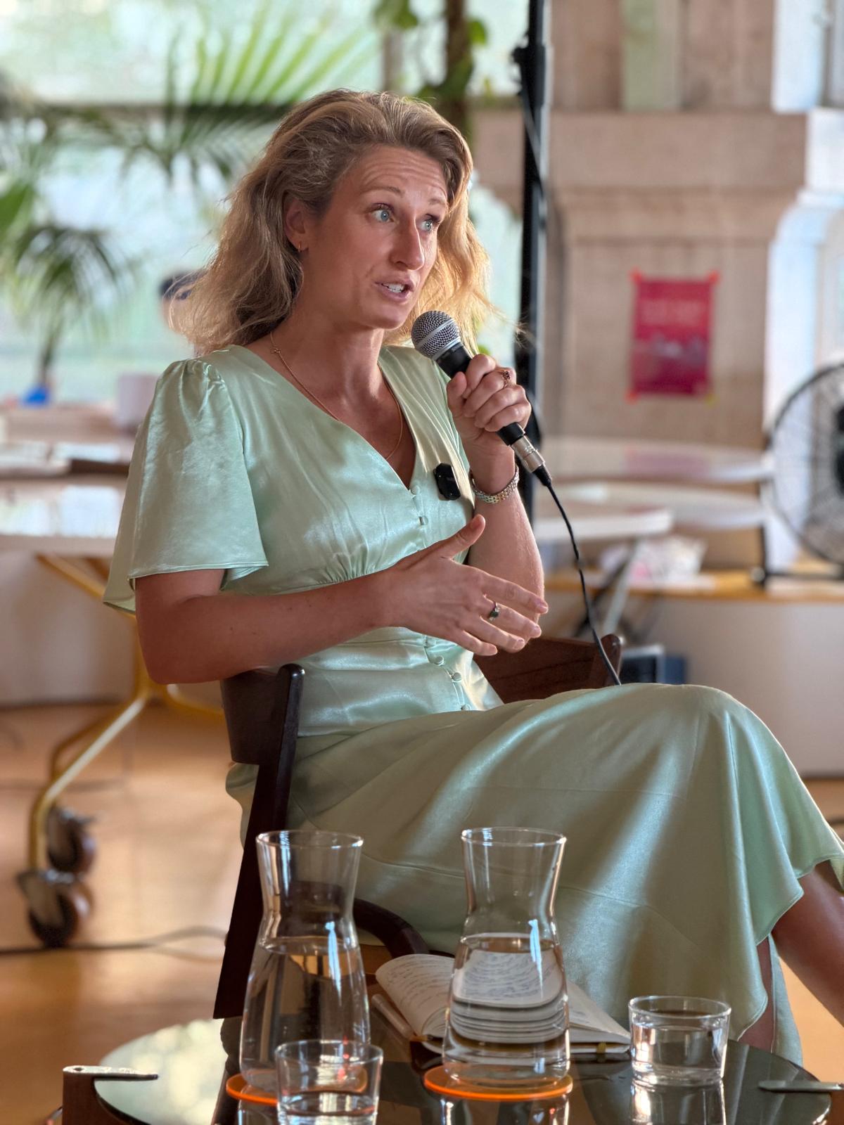 Ella Writer seated in an armchair, speaking into a handheld microphone and gesturing with an open hand